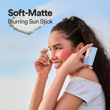 SaltTree model smiling while holding a TOCOBO Cotton Airy Sun Stick SPF 50 against a clear blue sky background.